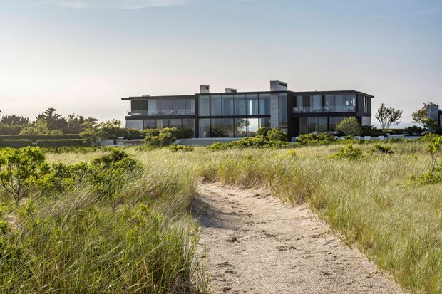 A large contemporary multi-storey residence with a dark, rectilinear facade, extensive floor-to-ceiling glazing, projecting upper-level forms, and flat rooflines. The house is viewed from a sandy path through coastal dune vegetation, with landscaped planting around the building and glimpses of water or horizon beyond.