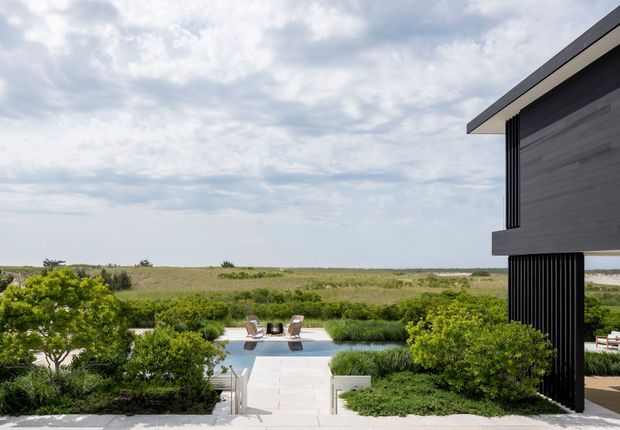 Exterior view of a contemporary dark-clad house beside a landscaped pool terrace, with a pale paved path leading through dense coastal planting toward a broad open landscape and water beyond. The building features strong horizontal lines, projecting roof planes, vertical black screening, and large glazed openings.