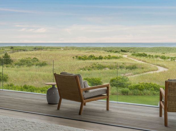 A tranquil coastal outdoor seating area viewed from a covered or glazed living space, with timber decking, woven timber armchairs, and a small side table overlooking grassy dunes, a sandy path, and the ocean horizon. The scene has a relaxed, natural resort-like feel with expansive glazing and an unobstructed coastal outlook.
