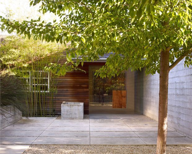 A landscaped residential courtyard or entry approach with large concrete paving slabs, a mature leafy tree, bamboo planting, and a compact modern timber-clad structure. The entrance combines horizontal dark wood cladding, a glazed door or window, a rust-toned metal panel, and a pale concrete block boundary wall. The image has a warm, tranquil, natural atmosphere with strong indoor-outdoor architectural detailing.