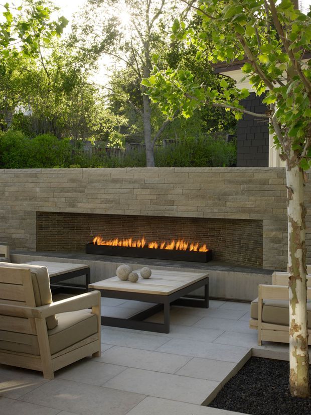A landscaped outdoor patio with a built-in linear gas fireplace set into a textured stone-clad wall. The space includes pale paved flooring, timber-framed upholstered lounge chairs, a low coffee table, mature trees, and dense garden planting, creating a refined outdoor entertaining area.