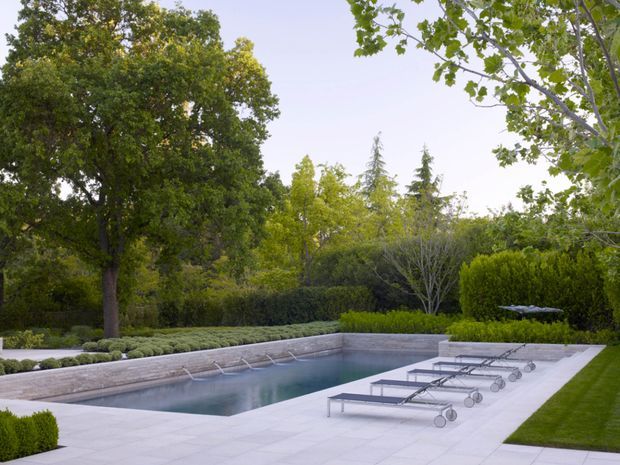 A landscaped residential pool area with a long rectangular swimming pool, pale stone or concrete paving, several wheeled loungers, clipped hedges, mature trees, and manicured lawn. The setting is calm and refined, with a clean contemporary garden design.