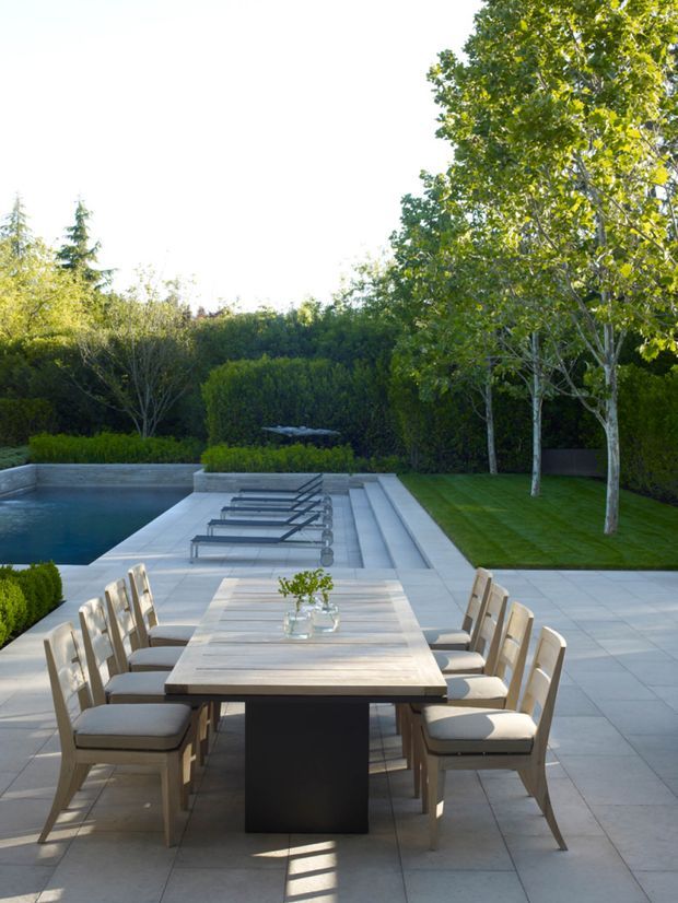 A landscaped outdoor dining terrace with a long timber dining table and upholstered chairs arranged beside a rectangular swimming pool. Pale stone or concrete paving extends around the pool, with broad steps, sun loungers, manicured hedges, lawn, and mature trees creating a refined garden setting.