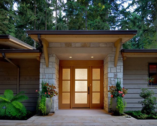 A covered residential entry porch set within a wooded landscape. The entrance features a warm timber-framed glazed double door, substantial pale natural-stone pillars and surround, exposed timber soffit, and dark horizontal exterior cladding. Symmetrical planters and lush ferns frame the paved approach, creating a sheltered, natural woodland entry.