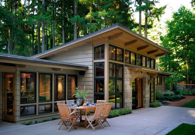 A contemporary single-storey house set within a dense forest, photographed from an outdoor patio. The home has broad low-pitched roof planes with deep timber-lined eaves, extensive dark-framed glazing, horizontal light-coloured wall cladding, and a prominent stone-clad entry column. A timber outdoor dining setting sits on a large paved terrace, with garden planting around the edges.