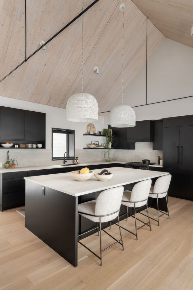 A contemporary kitchen dominated by a large island with a pale stone benchtop, dark matte cabinetry, and four upholstered bar stools. The kitchen has light timber flooring, a light timber-lined pitched ceiling with exposed black structural detailing, a pale textured backsplash, open shelving, and two oversized woven pendant lights. The overall palette is warm and neutral with strong black-and-white contrast.
