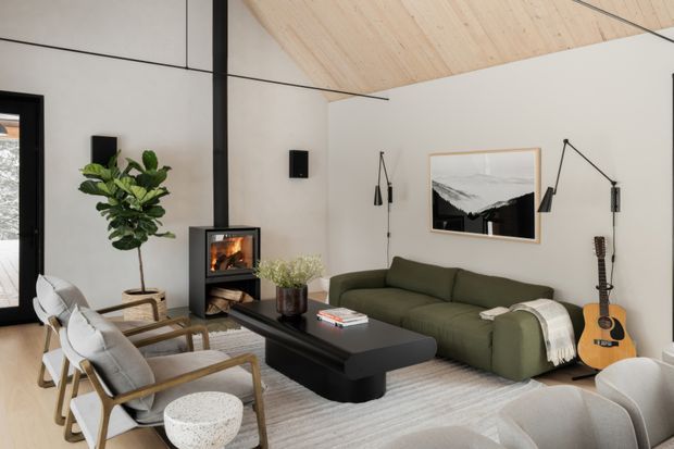 A bright contemporary living room with a pitched light timber-lined ceiling, pale walls, black-framed glazing, and a black freestanding fireplace. The room is furnished with an olive-green sofa, light upholstered timber-framed lounge chairs, a black coffee table, a pale rug, floor lamps, indoor greenery, artwork, and a guitar. The space has a warm, refined, nature-inspired character with strong contrast between light finishes and dark architectural accents.