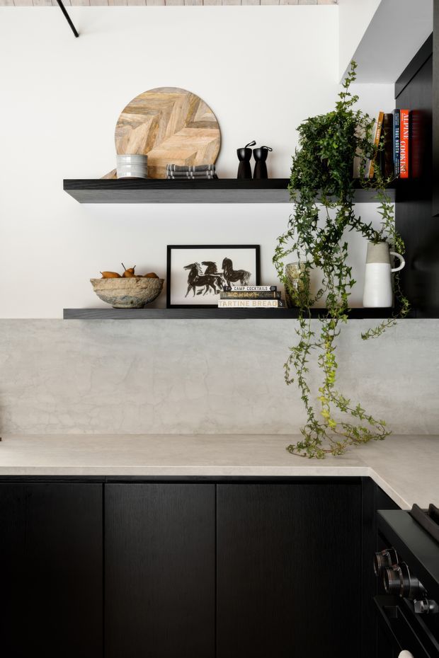 A close-up view of a sophisticated contemporary kitchen. Matte black lower cabinetry and a dark benchtop frame a pale grey stone or concrete-look splashback and benchtop surface. Two slim black display shelves hold decorative ceramics, books, artwork, and a trailing indoor plant. The composition is monochromatic with warm timber accents and refined styling.