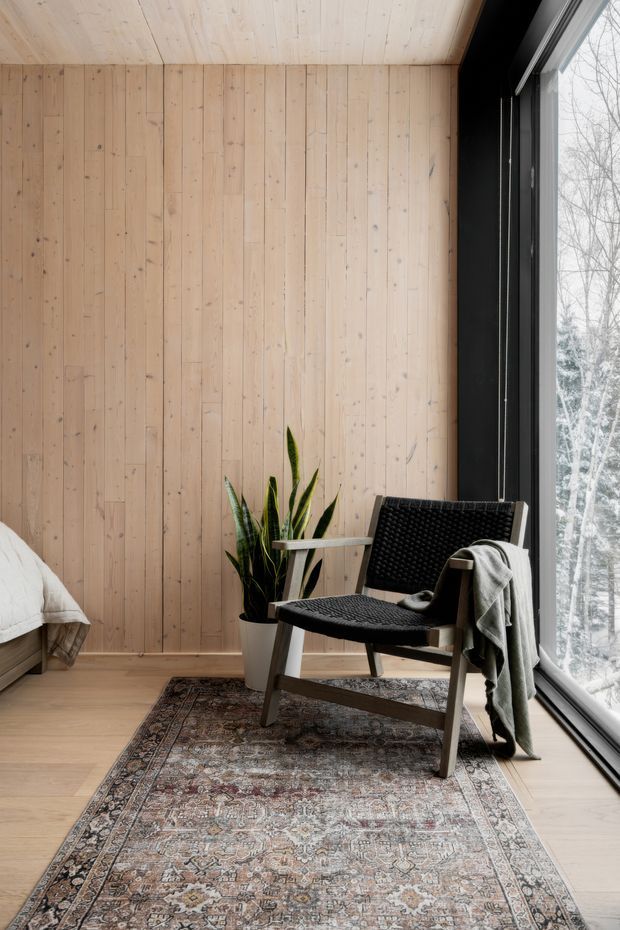 A serene, minimalist reading corner in a wood-lined room. Pale vertical timber wall and ceiling boards meet light timber flooring, with a large black-framed floor-to-ceiling window overlooking trees. A woven black lounge chair, potted plant, draped grey throw, patterned vintage rug, and the edge of a bed create a warm, natural retreat.