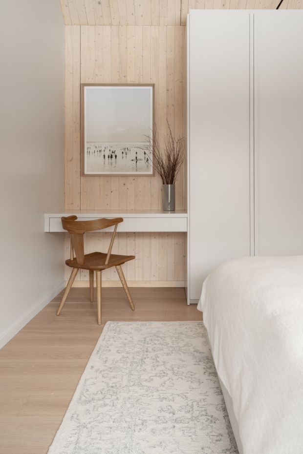 A softly lit, minimalist bedroom corner featuring a white bed, pale timber flooring, a full-height white wardrobe, and a compact floating desk against a light vertical timber-panelled wall. A sculptural wooden chair, framed beach photograph, and tall vase with branches add restrained natural detail. The palette is warm, neutral, and understated.