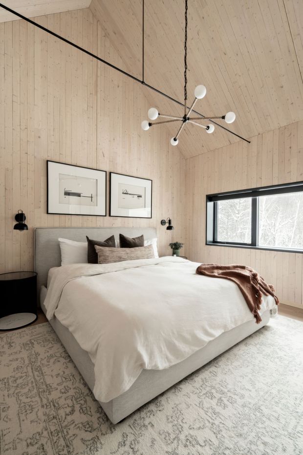 A bright, contemporary bedroom with a large upholstered bed dressed in white bedding and layered neutral pillows. Pale vertical timber wall and ceiling panelling create a warm, natural envelope, complemented by black-framed glazing, minimalist black bedside tables and sconces, framed artwork, a sculptural multi-arm pendant light, and a large muted patterned rug. Snowy trees are visible through the window.