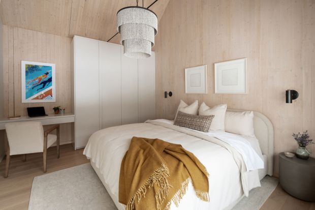 A warm, compact contemporary bedroom with pale timber-lined walls and ceiling, a white upholstered bed dressed in layered white bedding, and a mustard throw. A full-height white wardrobe occupies the rear wall, with a small built-in desk and chair beneath colourful artwork at left. Black wall sconces, framed artwork, a woven tiered pendant, and a rounded bedside table add restrained decorative detail. The room has a calm, refined, natural feel with soft neutral tones and timber surfaces.