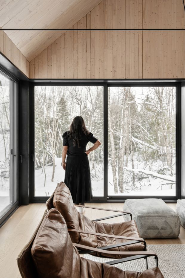 A warm, modern living room with full-height black-framed glazing overlooking a snowy wooded landscape. The room features pale timber flooring, a dramatic sloped timber-lined ceiling and wall, leather lounge chairs with black metal frames, and a large textured upholstered ottoman. A person stands by the windows, emphasizing the connection to the winter setting.