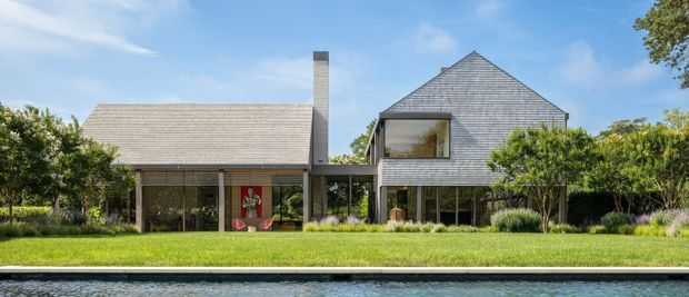 Front exterior view of a refined two-part residential property beside a swimming pool. The home combines a low-pitched gabled wing with a taller steeply gabled volume, both clad in grey weathered-looking shingles or tiles, with extensive glazing, a covered link, landscaped garden, and mature hedging.