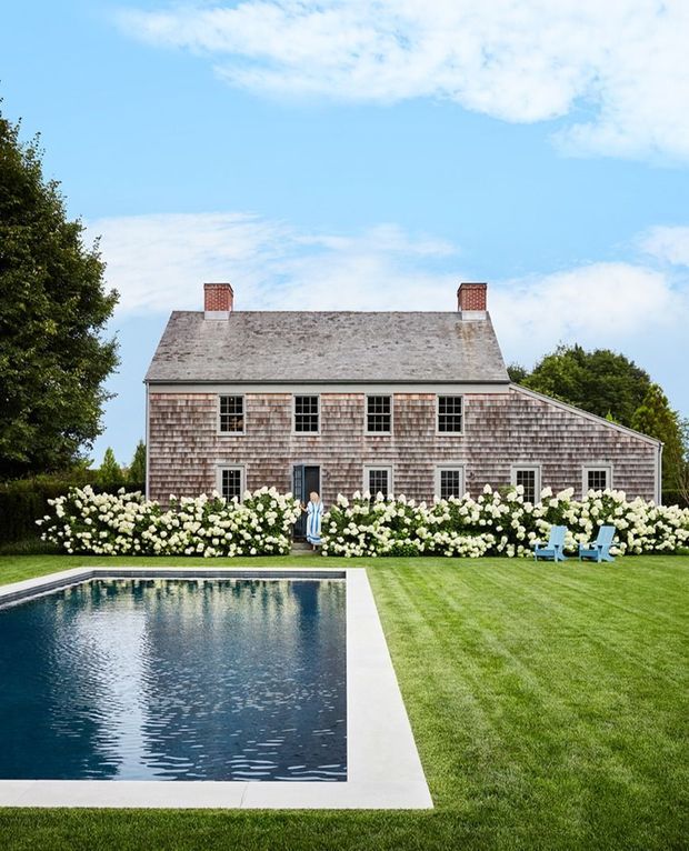 A formal landscaped backyard featuring a large rectangular swimming pool with pale paved edging, manicured lawn, flowering shrubs, and blue Adirondack-style chairs. Beyond the garden is a traditional shingle-clad house with a steep gabled roof, two brick chimneys, symmetrical multi-pane windows, and a long side extension. The scene is bright and summery with a calm, refined residential atmosphere.