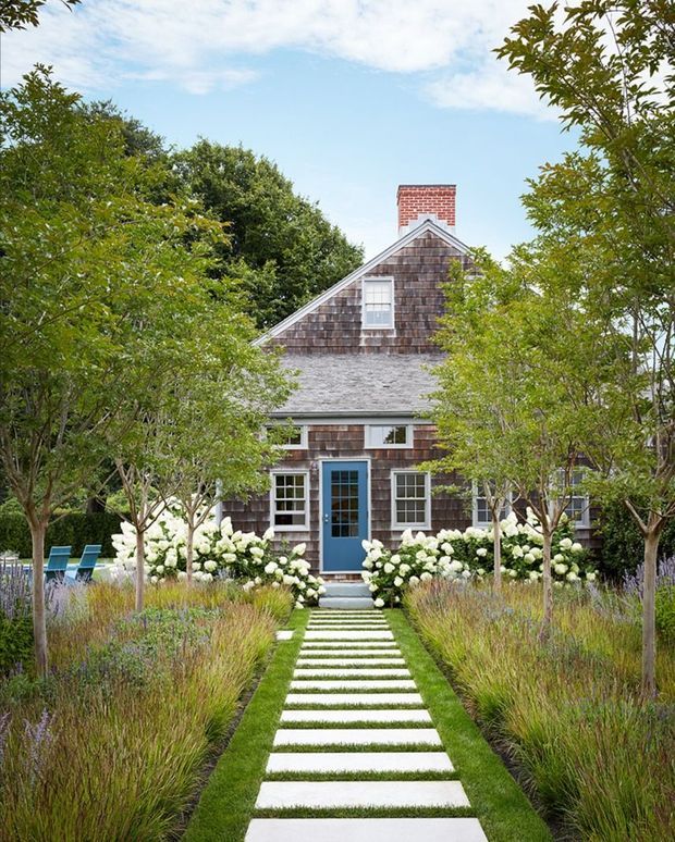 Front exterior view of a charming shingle-clad cottage partially framed by mature trees and landscaped planting. A straight path of large pale stepping slabs through clipped lawn leads to a blue front door, with white-trimmed windows, a steep gabled roof, and a red brick chimney. Hydrangeas, ornamental grasses, and garden seating contribute to a refined cottage garden setting.