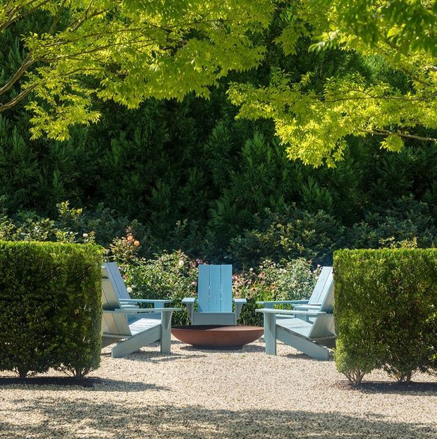 A manicured outdoor garden seating area with pale gravel paving, clipped hedge screens, lush layered planting, and several light-coloured lounge chairs arranged around a shallow circular fire bowl or planter. Mature tree canopy provides dappled shade, creating a serene formal garden atmosphere.