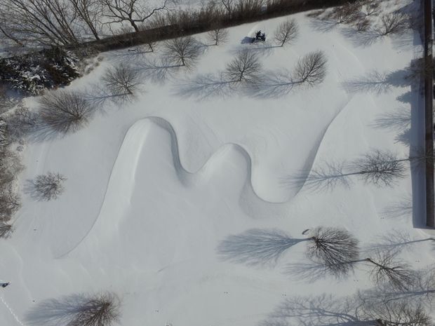 Aerial view of a snow-covered landscaped site with a sculpted, undulating snow-covered landform or roof-like surface, sparse leafless trees casting long shadows, and a dark linear building edge visible along the right side. The image is monochromatic and wintry, with no clearly identifiable interior space.