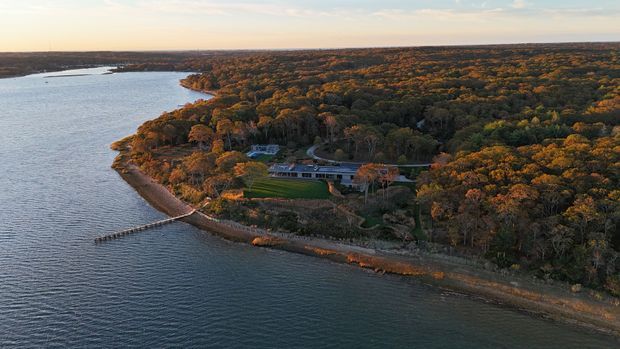 Aerial view of a secluded modern property set within dense autumn woodland along a broad waterfront. The long, low building sits near the shoreline, with landscaped grounds and a small private pier extending into the water.