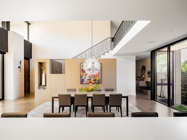 Bright contemporary open-plan dining area with a long rectangular dining table and upholstered chairs, positioned beneath a dramatic double-height void. A sculptural staircase with slim dark metal balustrading rises behind the dining space, alongside light timber and white built-in wall panels. Floor-to-ceiling glazing opens to an exterior terrace or garden, while abstract artwork and decorative objects add colour to the otherwise restrained interior.