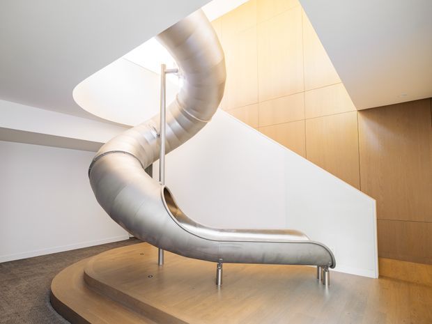 A sculptural indoor spiral slide descends through a bright, minimalist multi-level interior. The slide is formed from brushed metal tubing and supported by slim metal posts, set on a raised curved timber platform. White walls, a sculptural curved opening, warm timber wall panelling, and a carpeted floor create a contemporary architectural setting.