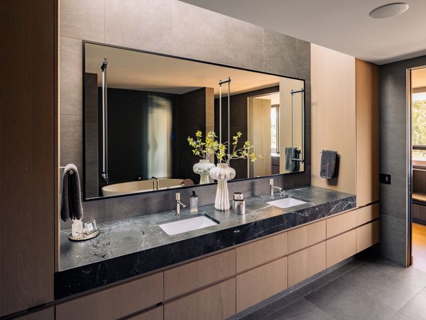 Contemporary double-vanity bathroom featuring a long dark stone benchtop, two integrated white basins, wall-mounted tapware, extensive mirrored cabinetry, warm timber veneer drawers, large grey wall and floor tiles, and black-framed detailing. A freestanding bath is visible in the mirror reflection, with a bedroom or adjoining room glimpsed through the doorway.