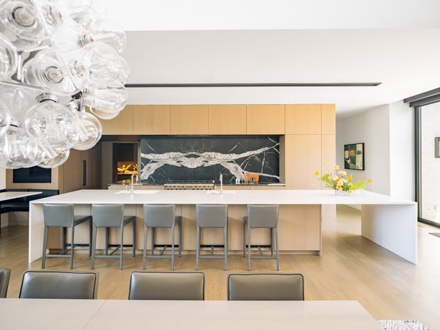Bright, spacious contemporary kitchen and dining area featuring a long white waterfall island with seating for six, pale timber cabinetry, light timber flooring, and a dramatic dark marble-look splashback with bold white veining. A sculptural clear-glass pendant light hangs over the dining area, with large windows and clean white walls contributing to an airy, refined interior.