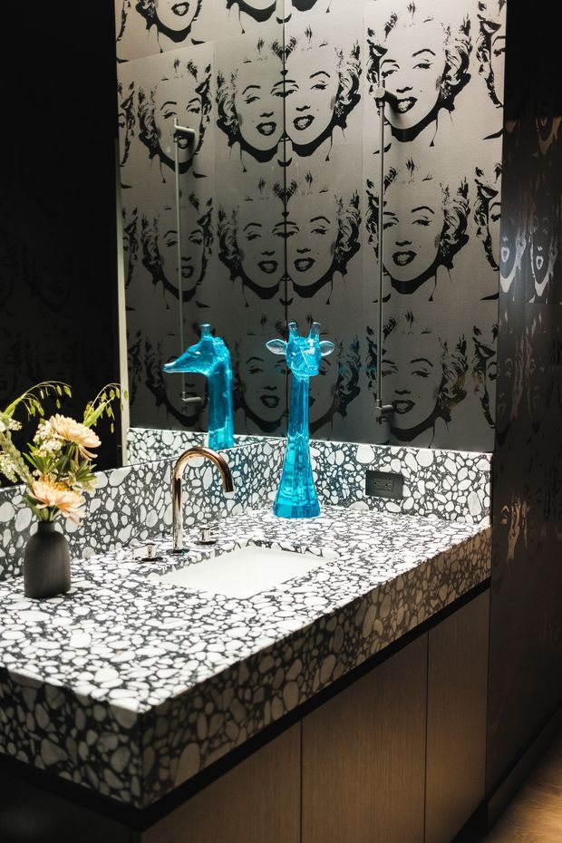 A dramatic powder-room vanity with a white sink set into a bold black-and-white patterned stone benchtop and matching splashback. The wall behind is covered in a repeated monochrome portrait motif, reflected in mirrored panels. Dark cabinetry, a brushed metal tap, and vivid turquoise sculptural glass vases create a theatrical, eclectic finish.