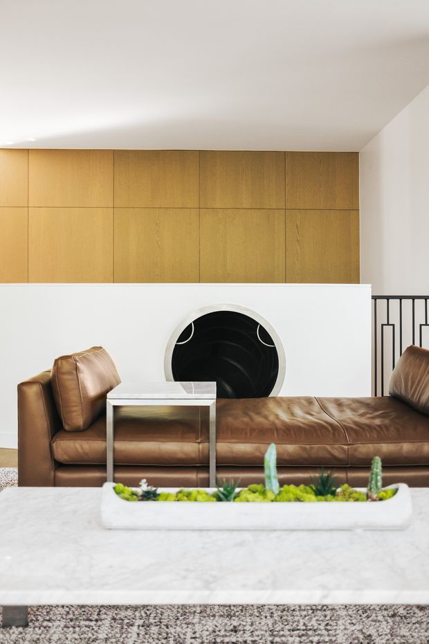 A refined contemporary living room featuring a large cognac-brown leather chaise sofa, a white marble coffee table, a low white planter with succulents, and a striking circular black wall feature set into a white partition. Warm wood-panelled cabinetry spans the upper wall, while a black geometric metal balustrade is visible at right. The room has a restrained neutral palette with warm timber and leather accents.