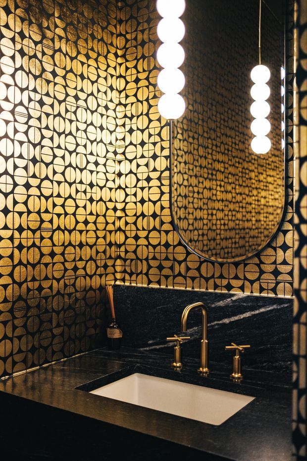 A dramatic, luxurious bathroom vanity featuring a black stone benchtop with an integrated white rectangular basin, brushed gold tapware, and a large rounded-rectangle mirror. The walls are covered in a highly distinctive black-and-gold geometric circular patterned tile, complemented by sculptural globe pendant lights reflected in the mirror. The space has a dark, richly layered palette and strong Art Deco-inspired detailing.