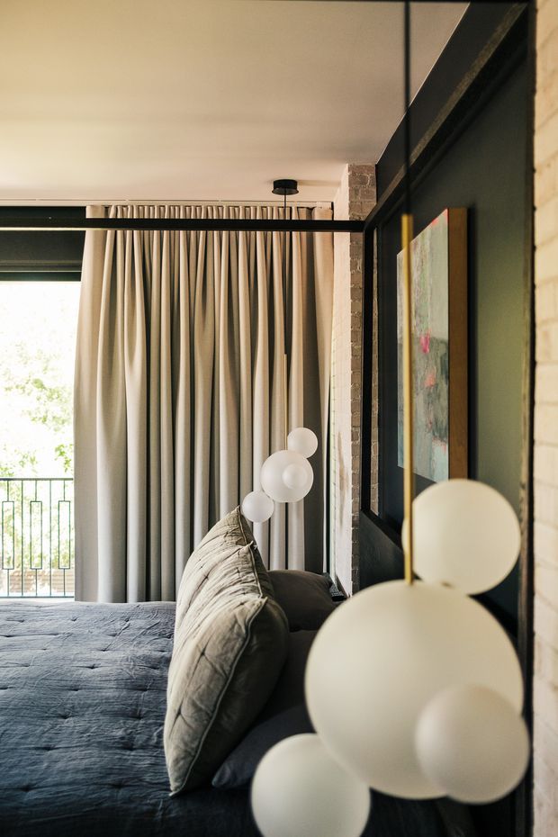 A warmly lit bedroom interior viewed from beside the bed. The room features a dark blue-gray upholstered bed with layered cushions, full-height pleated beige curtains, black-framed glazing or internal partitions, exposed brick detailing, and sculptural globe pendant lighting. A balcony and leafy exterior are visible beyond the curtains. The composition has a refined contemporary feel with strong black, cream, and charcoal contrasts.