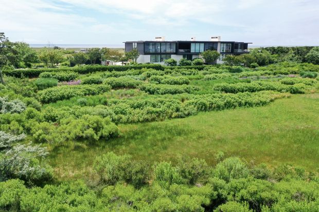 Wide exterior view of a low, elongated contemporary residence set within expansive landscaped gardens and dense coastal vegetation. The building has a dark linear facade, extensive floor-to-ceiling glazing, flat rooflines, and projecting wings. A broad water or ocean horizon is visible behind the property.