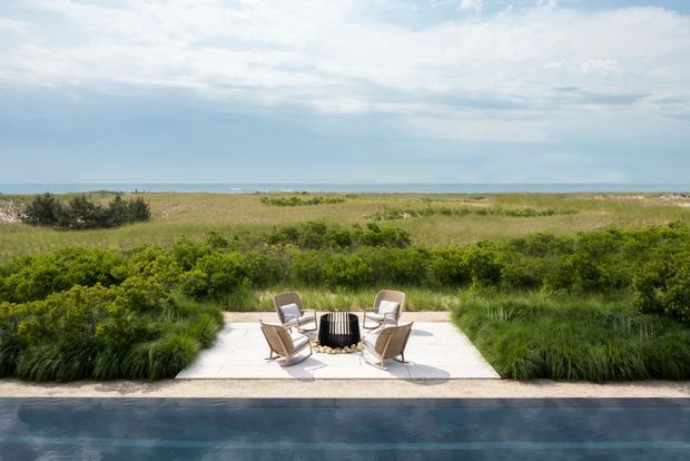 A landscaped outdoor terrace beside a swimming pool, furnished with four lounge chairs around a black outdoor fire pit. Dense ornamental grasses and coastal vegetation frame the pale paved seating area, with a broad ocean horizon visible beyond the dunes.