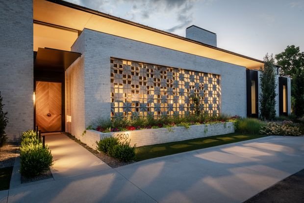 Exterior architectural photograph of a contemporary single-storey residence with a flat roof, white-painted brick walls, warm recessed lighting, and a prominent decorative breeze-block screen across the front facade. A timber entrance door sits beneath a covered entry, while dark vertical facade elements frame the right side. The landscaped frontage includes a raised planter and paved driveway or walkway.