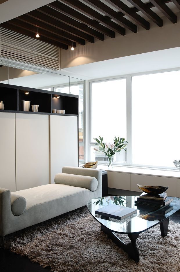 A refined contemporary living room with a pale upholstered chaise lounge, glass coffee table, shag rug, and minimalist decorative objects. Floor-to-ceiling windows provide abundant daylight, while dark exposed ceiling beams contrast with white walls and built-in cabinetry. A mirrored wall and integrated shelving add depth and visual interest.