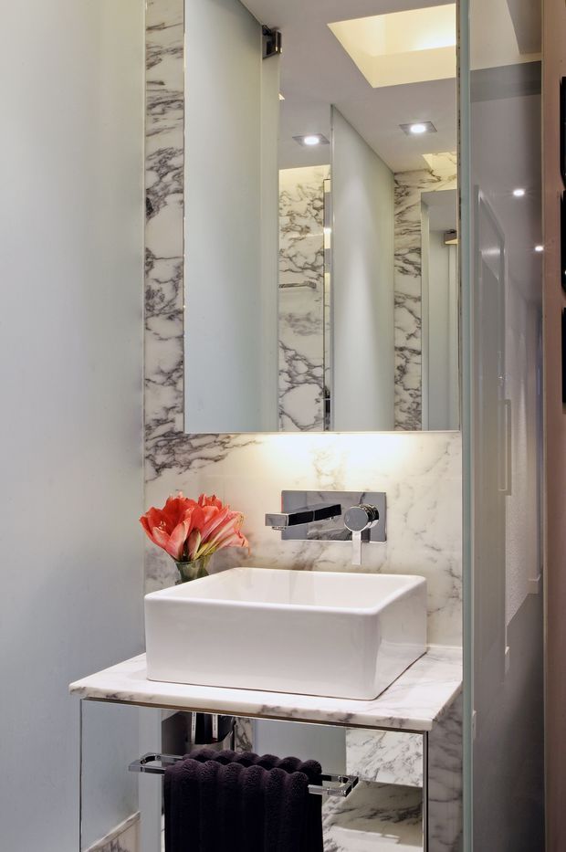 Compact contemporary bathroom vanity with a white vessel basin, wall-mounted chrome tapware, mirrored cabinet, marble-look wall and benchtop surfaces, and integrated lighting. A dark towel rail and small floral arrangement add contrast.