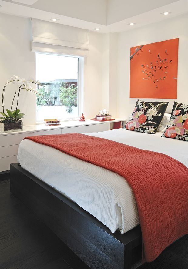 Bright contemporary bedroom with a low-profile dark built-in bed platform, crisp white bedding, and a textured terracotta-red bed throw. Floral-patterned black cushions add contrast, while a matching orange-red artwork provides a bold focal point. The room features white walls, recessed ceiling lighting, a large window with a Roman blind, dark flooring, and a built-in window seat or storage ledge decorated with books and an orchid.
