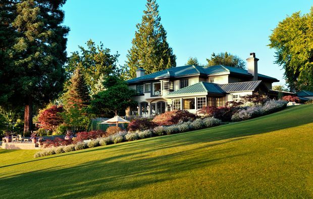 A large multi-level house set on a gently sloping landscaped lawn, surrounded by mature trees, layered ornamental planting, and outdoor seating. The home has a traditional-inspired facade with light-coloured cladding, extensive multi-pane windows, several intersecting pitched roofs with dark metal roofing, covered verandas, and prominent chimneys. The image is a bright exterior architectural view in a verdant suburban or garden setting.