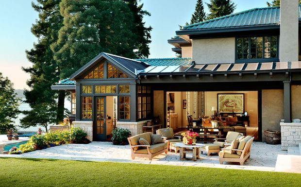 A large traditional-style residence viewed from the landscaped garden, featuring a covered outdoor living terrace with lounge seating, a stone-paved patio, extensive glazed doors and windows, and multiple blue-green tiled pitched roofs. The house combines pale rendered walls, dark timber or metal framing, and a glazed gabled sunroom. Mature trees and a glimpse of water appear in the background.