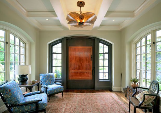 Elegant, symmetrical sitting room or entry hall with a richly paneled central timber door framed by dark arched glazing. Large arched multi-pane windows line both sides, bringing in abundant daylight. The room features a coffered ceiling, decorative butterfly-like pendant light, pale green walls, patterned blue upholstered armchairs, a console table, and a large muted red patterned rug.