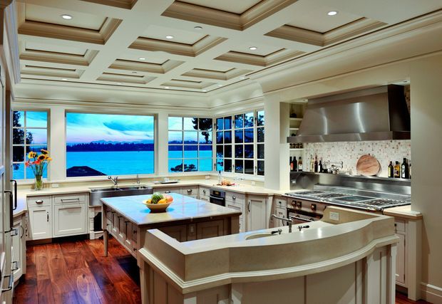 A spacious, traditionally styled kitchen with extensive white cabinetry, a large central island, a curved raised breakfast bar, and stainless-steel cooking appliances. The room features dark polished timber flooring, a tiled backsplash, and large multi-pane windows overlooking a bright waterfront landscape. The ceiling has elaborate coffered detailing with recessed lighting.