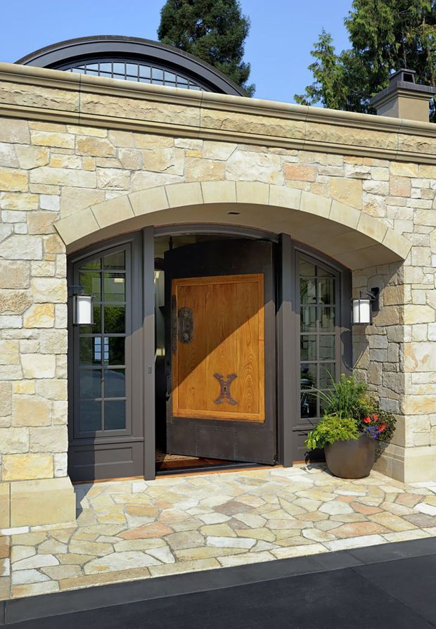Exterior view of a formal stone-clad entry facade. A large arched masonry opening frames a dark timber front door with decorative metal hardware, flanked by glazed sidelights and wall lanterns. The facade has substantial stonework, a projecting cornice, and a curved glazed roof or clerestory element above. Stone paving leads to the entry, with a potted plant beside the door.
