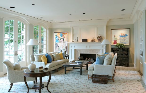 Bright, refined living room with pale blue-green walls, extensive white architectural trim, tall arched multi-pane windows, a decorative fireplace mantel, and symmetrical built-in detailing. The room is furnished with neutral upholstered sofas and armchairs accented by blue and yellow cushions, a dark coffee table, side tables, artwork, and a patterned carpet. The overall character is traditional with a polished, classic interior scheme.