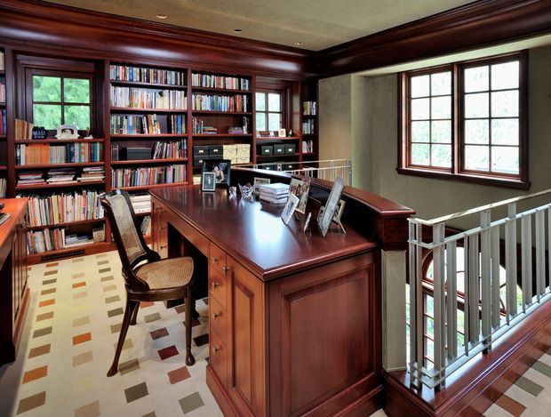 A richly detailed traditional home office or study occupying an upper-level landing. Dark stained wood built-in bookcases line the walls, complemented by a large matching central desk, filing drawers, classic trim, and framed multi-pane windows. A patterned carpet, wooden chair, framed photographs, and a white balustrade overlooking the level below complete the space.