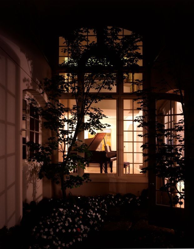 A dark, atmospheric view into a landscaped courtyard through large multi-pane windows and glazed doors. Warm interior lighting reveals a grand piano inside, while silhouetted trees and planting frame the facade. The image emphasizes the relationship between an intimate garden courtyard and the illuminated interior at night.