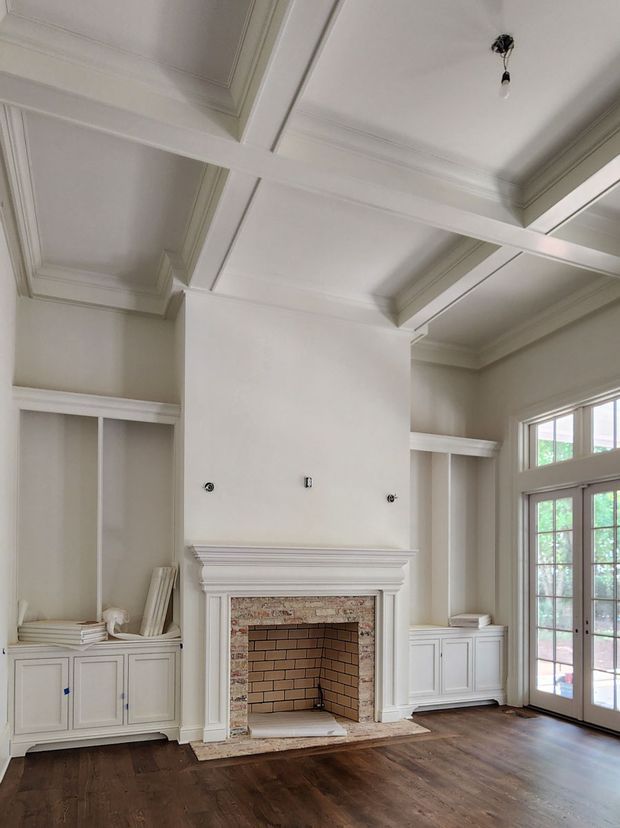 Bright, largely unfurnished traditional interior centered on a white-painted fireplace with a brick firebox and stone or brick surround. Built-in cabinetry and open shelving flank the fireplace, while tall French doors with divided-light glazing bring in natural light. The room has a dark timber floor, ornate ceiling coffers and beams, and substantial classical mouldings.
