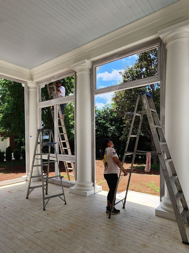 A bright enclosed porch or verandah undergoing window installation or renovation. The space has white-painted timber floorboards, a white beadboard ceiling, large glazed openings, and prominent classical white columns with decorative capitals and bases. Workers and ladders are visible, with a landscaped garden beyond the windows.