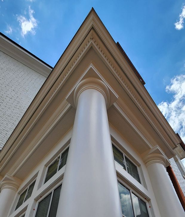 Low-angle close-up of an ornate multi-storey building facade, dominated by large smooth white columns supporting a projecting triangular pediment and richly detailed classical cornices. White-painted brick walls, multi-pane windows, decorative soffit detailing, and a bright blue sky are visible. The architecture has a formal traditional character.