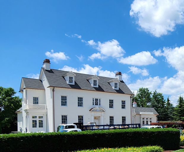 Exterior view of a large symmetrical white multi-storey residence with a steep dark grey slate-like gabled roof, multiple dormer windows, chimneys, classical window proportions, and a small portico entrance. The property is framed by manicured hedges and mature trees under a bright blue sky.