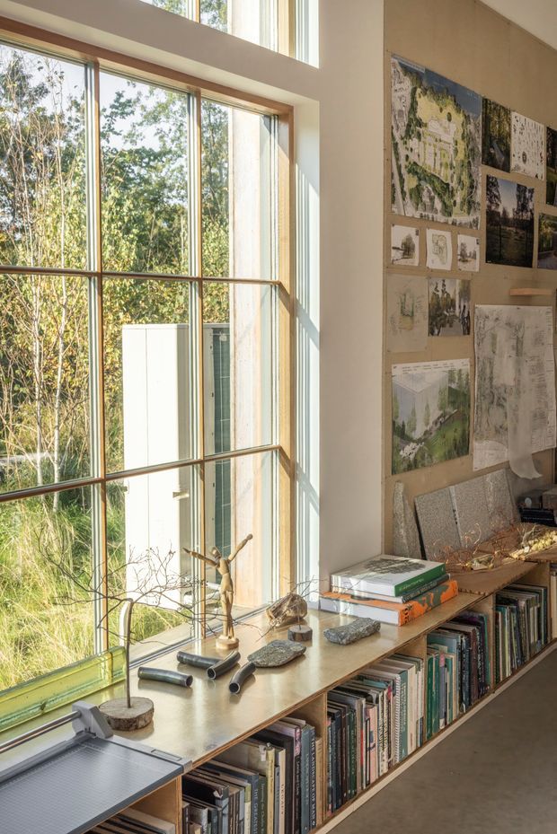 A warm, light-filled study or design workspace with a long built-in timber shelving unit and desk beneath tall multi-pane windows. Books, architectural drawings, maps, models, stones, and design materials are arranged along the work surface and displayed on the wall. The room has a calm, creative studio atmosphere with abundant greenery visible outside.